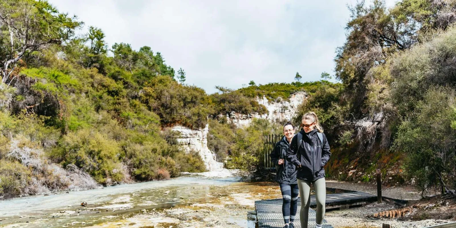 Two visitors walking the boardwalk through Wai-O-Tapu's pale sinter valley